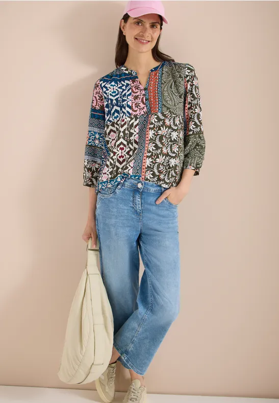 Frau vor beigem Hintergrund trägt bunte Patchwork-Bluse, weite Jeans und weiße Tasche.
