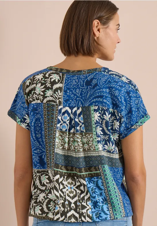 Rückansicht einer Frau, die ein kurzärmeliges Oberteil mit blau-grünem Patchwork aus floralen und geometrischen Mustern trägt.