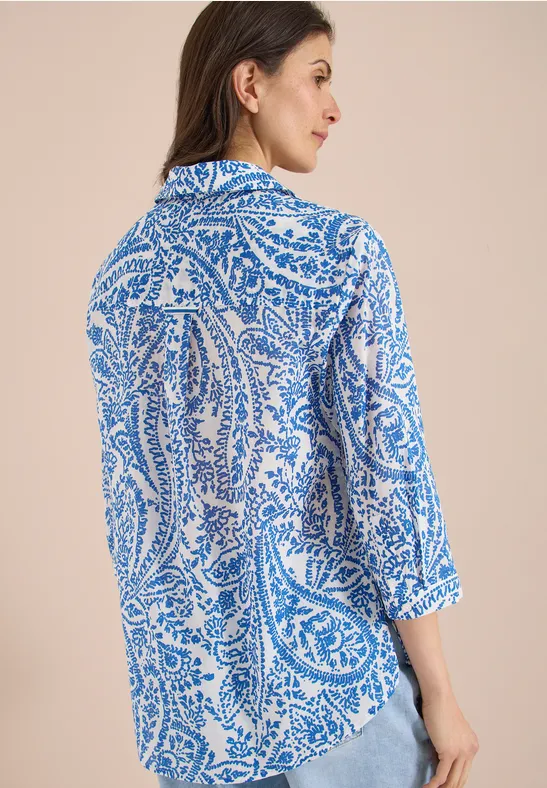 Rückenansicht einer Frau in Bluse mit blau-weißem Paisley-Muster und ¾-Ärmeln vor neutralem Hintergrund.
