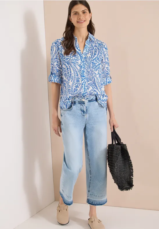 Frau lehnt an Wand, trägt blau gemusterte Bluse, weite Jeans, bequeme Schuhe und schwarzen Flechtkorb.