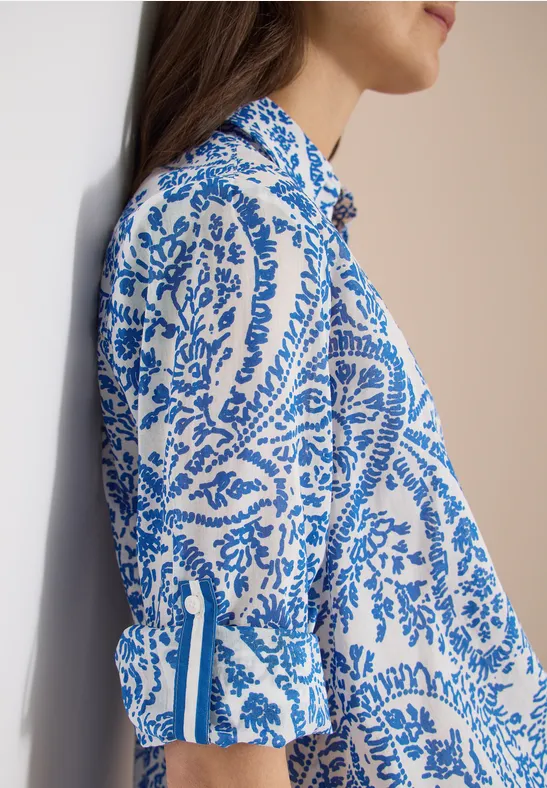 Seitliche Ansicht einer Frau in weißer Bluse mit blauem Paisley-Muster, am Wandrand lehnend.