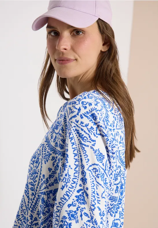 Eine seitlich abgewandte Person trägt ein weiß-blau gemustertes Oberteil und eine rosa Cap vor hellem Hintergrund.