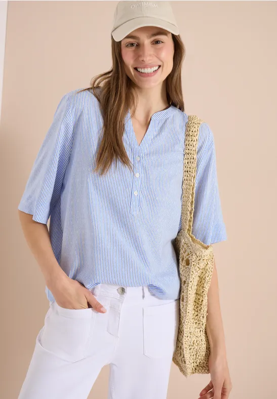 Vrouw in lichtblauwe 3/4 mouw blouse, witte broek en strooien tas tegen een neutrale achtergrond.