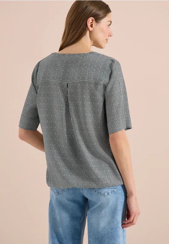 Frau von hinten in grauem gemustertem Oberteil mit halblangen Ärmeln und blauer Jeans.