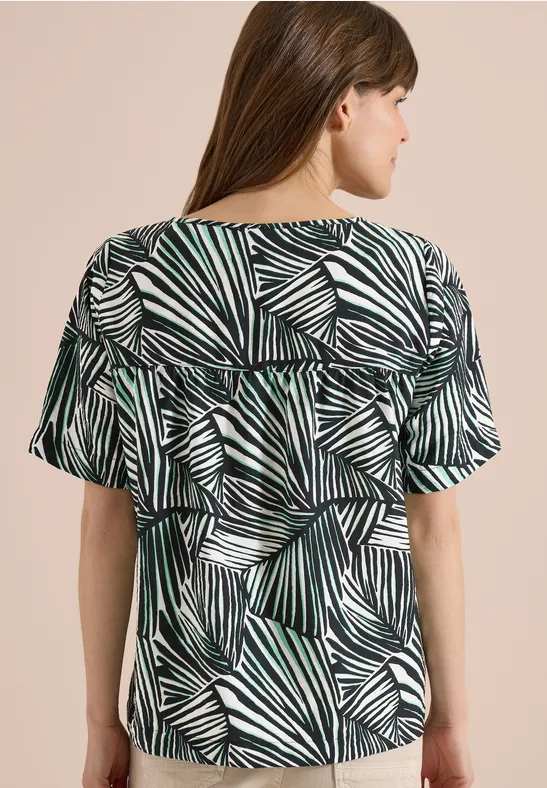 Frau von hinten in kurzärmeliger Bluse mit grün-weiß-schwarzem Blattmuster.