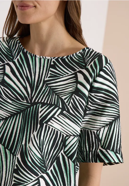 Frau in Top mit grünem und schwarzem Blattmuster und kurzen, leicht ausgestellten Ärmeln.