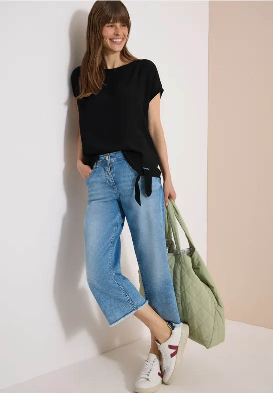 Frau in schwarzem T-Shirt, weitem Jeans-Culotte, weiß-roten Sneakern und grünem Stoffbeutel vor hellem Hintergrund.