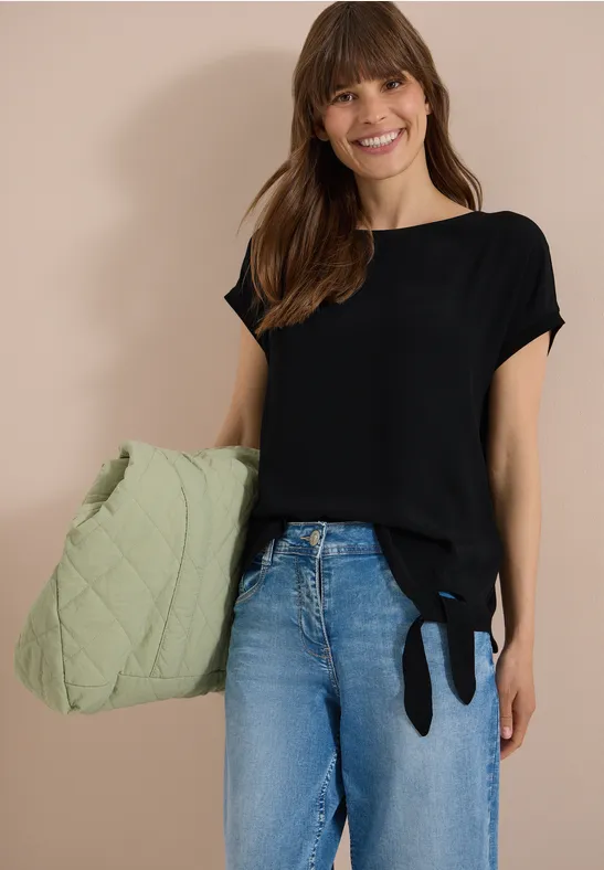 Frau in schwarzem Shirt mit Bändchen-Detail, blauer Jeans und großer gesteppter grüner Tasche.