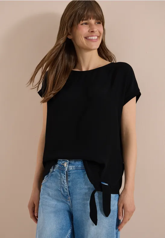 Eine Frau trägt eine schwarze Bluse mit seitlichem Knoten und blaue Jeans.