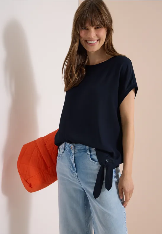 Frau in dunkelblauem, seitlich geknotetem T-Shirt, hellen Jeans, roter Tasche lässig unter dem Arm.