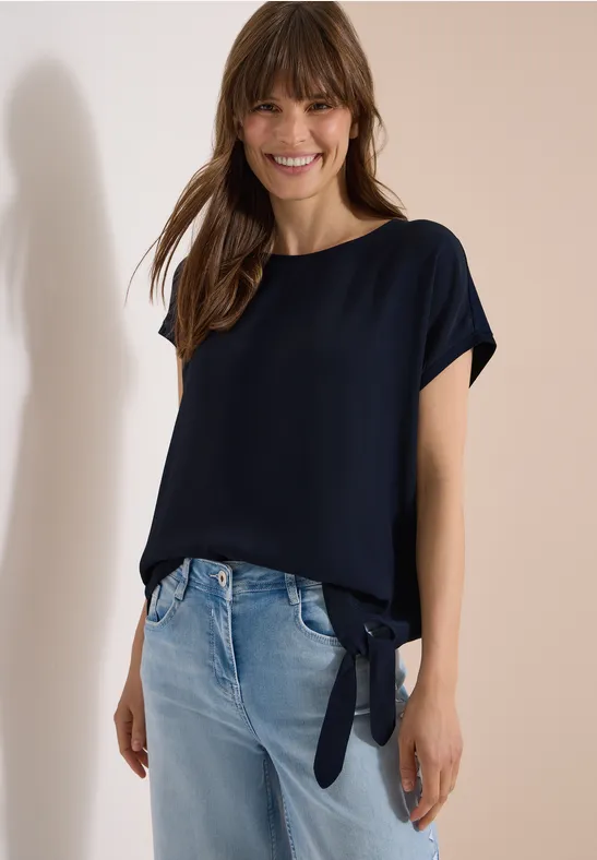 Frau in dunkelblauer, locker sitzender Bluse mit Seitenschlitz und Knoten, dazu helle Jeans vor hellem Hintergrund.