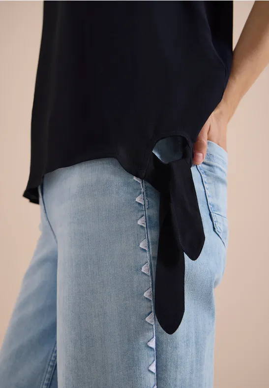 Schwarzes Top und helle Jeans mit seitlichem Dreieckbesatz, Hand in der Tasche.