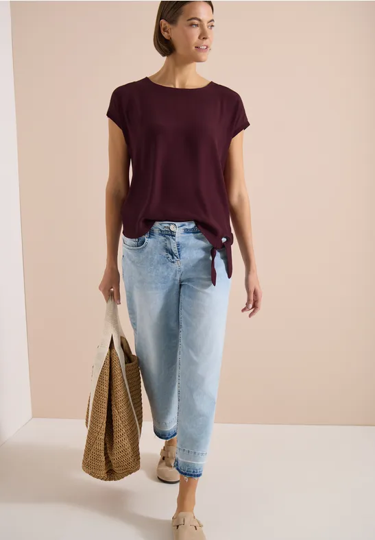 Frau in burgunderfarbenem T-Shirt und hellen Jeans mit Strohtasche in neutralem Raum.