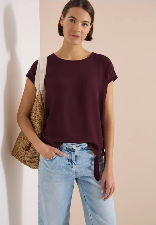 Person in lila T-Shirt, blauen Jeans und Strohtasche steht lässig vor weißer Wand.