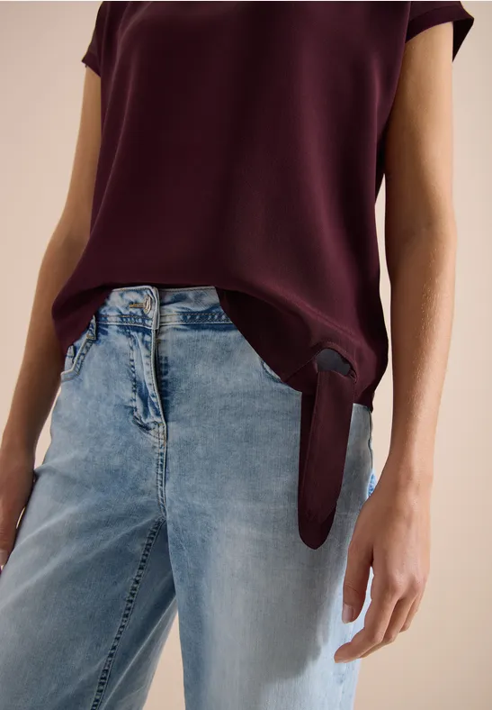 Frau trägt weinrote Bluse, vorne locker gesteckt, zusammen mit weiten hellblauen Jeans.