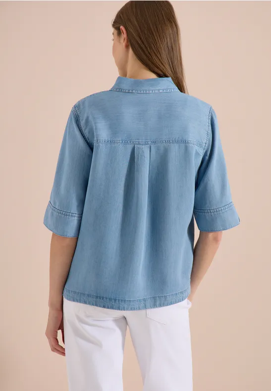 Achteraanzicht van een vrouw in een lichtblauwe spijkerblouse en witte broek.