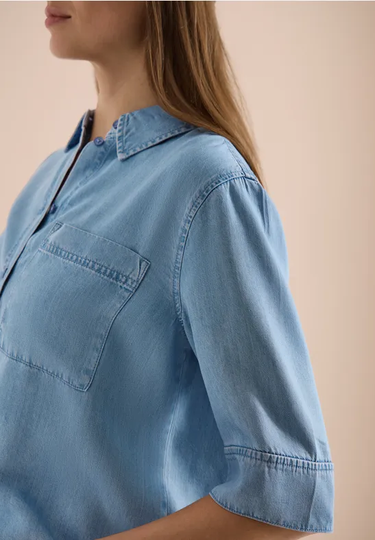 Zijaanzicht van een vrouw in een lichtblauwe denimblouse met borstzak en halflange mouwen tegen een beige achtergrond.