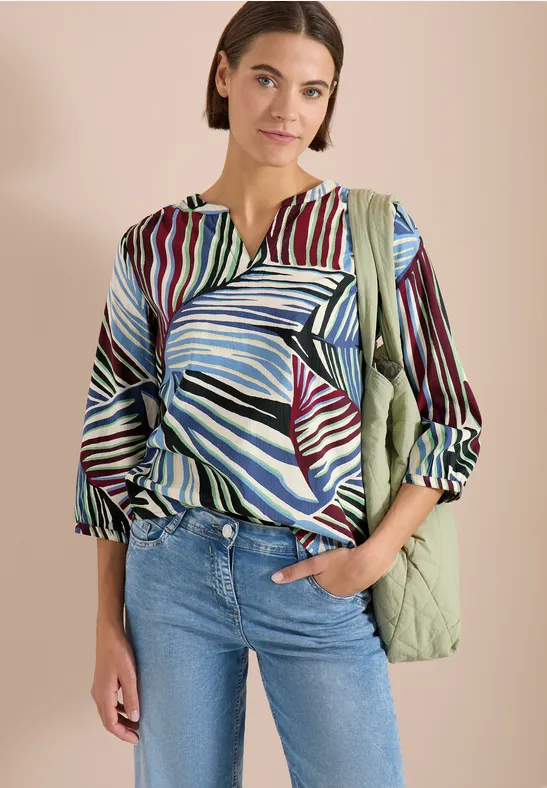 Vrouw met kleurrijk blouse met bladerenprint, blauwe jeans en lichtgroene gewatteerde tas voor beige achtergrond.