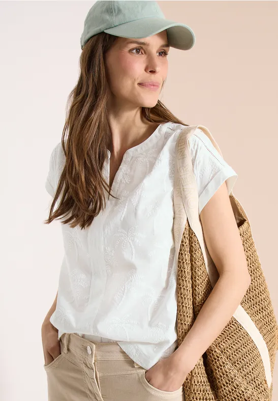 Eine Frau trägt weiße Bluse, beigefarbene Hose, Cap und eine große geflochtene Tasche.