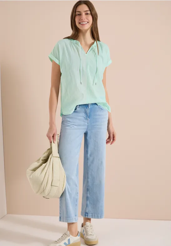 Femme en blouse menthe, jean large, baskets blanches et sac vert clair.