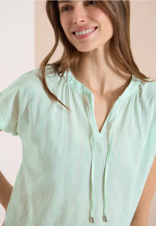 Femme portant un chemisier vert menthe à manches courtes, fines rayures blanches et col en V avec cordon.