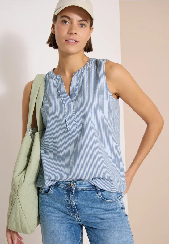 Femme en débardeur bleu clair, jean, sac vert à l’épaule et casquette blanche.
