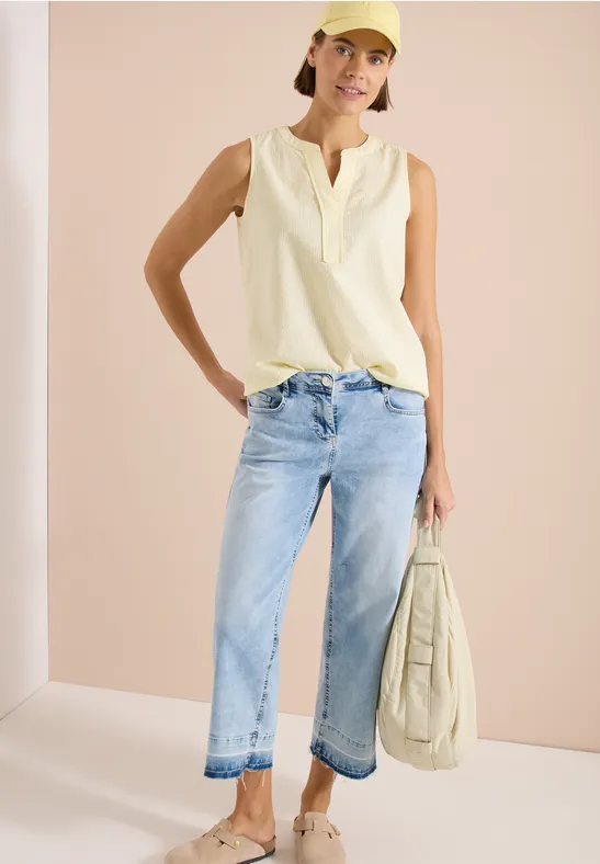 Frau in ärmellosem cremefarbenem Top, hellblauen Jeans und passender Tasche vor neutralem Hintergrund.