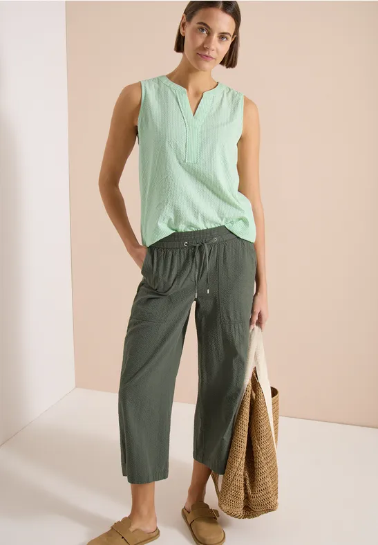 Femme en top menthe sans manches, pantalon kaki court, sandales fines et sac en osier.