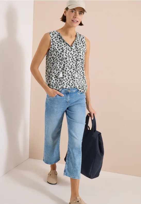 Vrouw met mouwloze top met print, wijde cropped jeans, beige loafers en zwarte tas tegen lichte achtergrond.