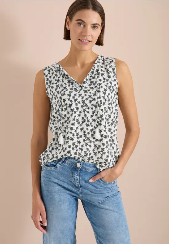Vrouw in mouwloze witte top met groen motief en V-hals, gecombineerd met een blauwe spijkerbroek.