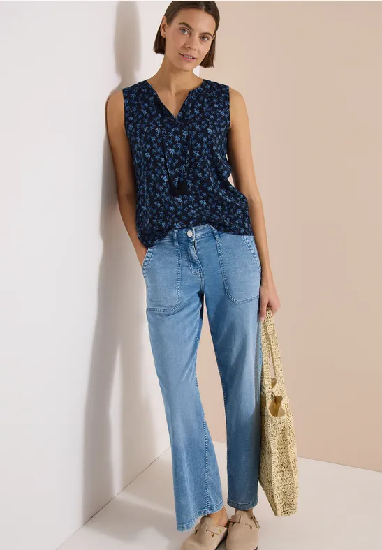 Femme portant un top bleu à fleurs sans manches, un jean clair et un sac en paille devant un fond bicolore.
