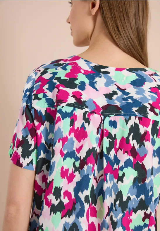 Achteraanzicht van een vrouw met een kleurrijke blouse met korte mouwen en een abstract patroon in roze, blauw, groen en wit.