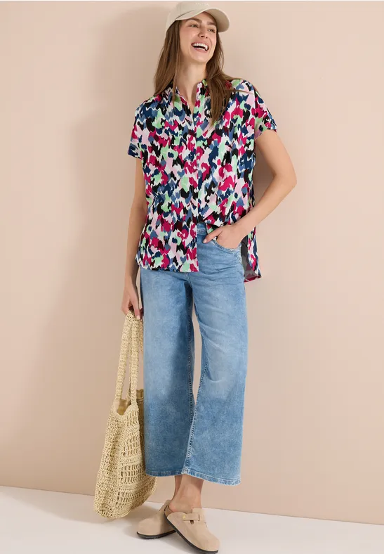 Eine Frau trägt eine bunte Kurzarmbluse, vorn geknotet, weite Jeans, Basttasche und Slipper.