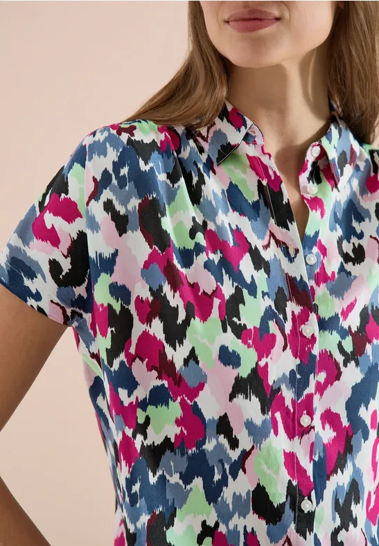 Frau trägt kurzärmliges Poloshirt mit buntem abstraktem Muster in Pink, Grün, Schwarz und Blau.