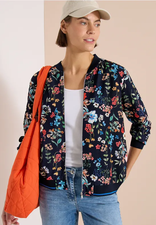 Eine Frau trägt eine schwarze Bomberjacke mit buntem Blumenmuster, Jeans, weißes Top, Cap und orangefarbene Tasche.