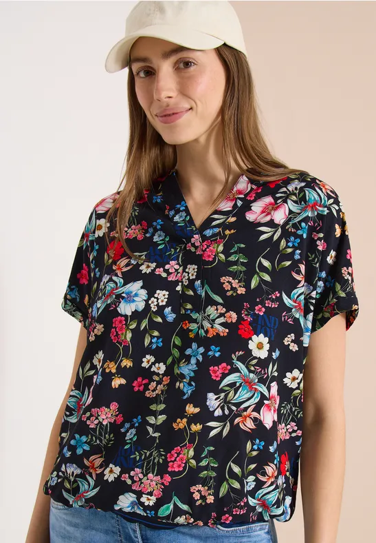 Frau vor hellem Hintergrund trägt schwarze, kurze Blumenbluse und helle Baseballkappe.