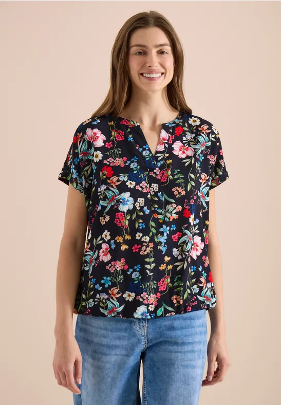 Frau in schwarzer Bluse mit buntem Blumenmuster und blauen Jeans, frontal stehend.