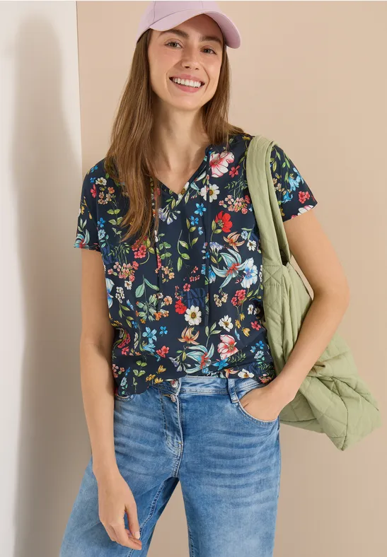 Frau in Jeans, dunkelblauer Bluse mit bunten Blumen, grünem Schulterbeutel, eine Hand in der Hosentasche.