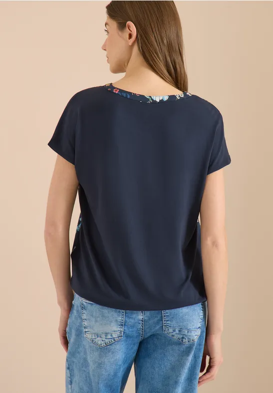 Frau von hinten in navyblauer Kurzarmbluse mit Blumenmuster am Kragen und hellen Jeans.