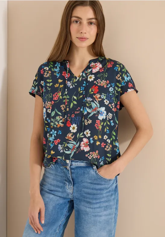 Frau in dunkelblauer, kurzärmeliger Bluse mit buntem Blumenmuster und Jeans, Hände in den Taschen.