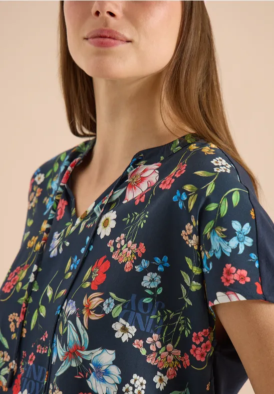 Frau trägt dunkelblaues Oberteil mit buntem Blumenmuster, V-Ausschnitt und kurzen Ärmeln.