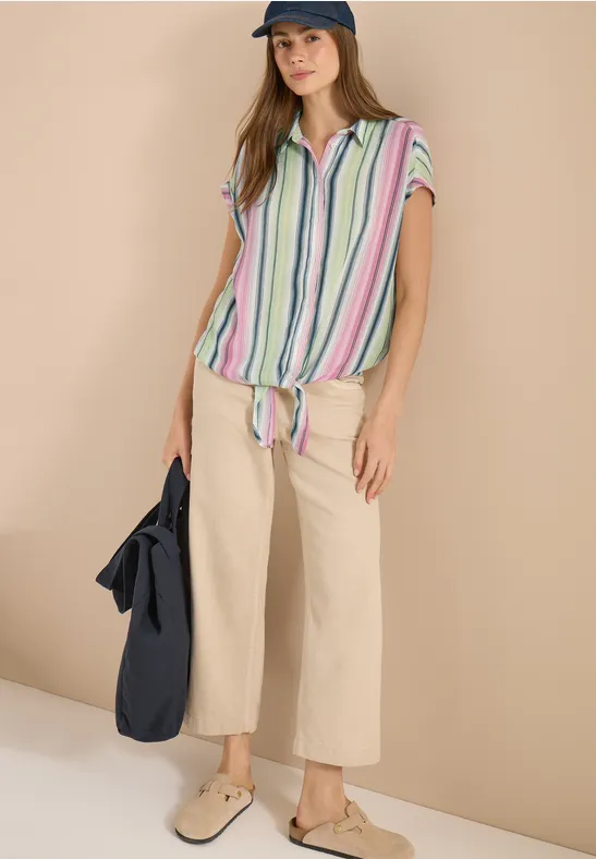 Persoon draagt beige wijde broek en gestreept geknoopt shirt, met tas en platte schoenen.