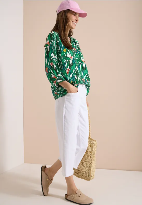 Eine Frau in grüner Blumenbluse, weißen Hosen und rosa Cap, mit Strohtasche und Mokassins.