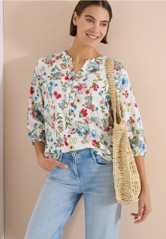 Femme en chemisier fleuri, jean clair et sac en paille tressée.