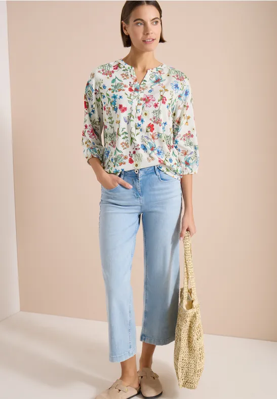 Femme portant un chemisier fleuri, jean bleu clair évasé, espadrilles et un sac en paille sur fond neutre.