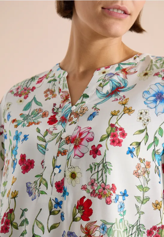 Femme porte un chemisier blanc à motifs floraux colorés et col en V.