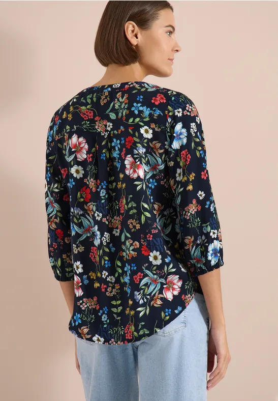 Femme de dos portant un chemisier bleu foncé à fleurs colorées, manches 3/4, et jean clair.