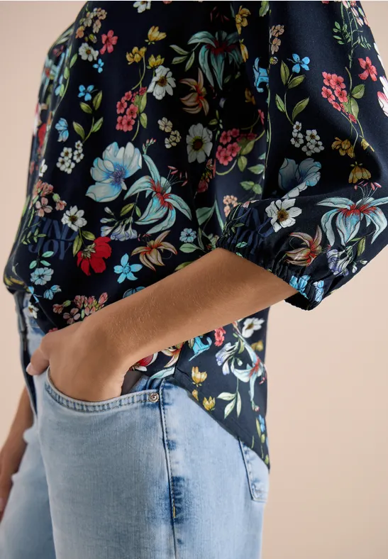 Haut femme à manches mi-longues et imprimé fleuri coloré, partiellement rentré dans un jean bleu clair, main en poche.