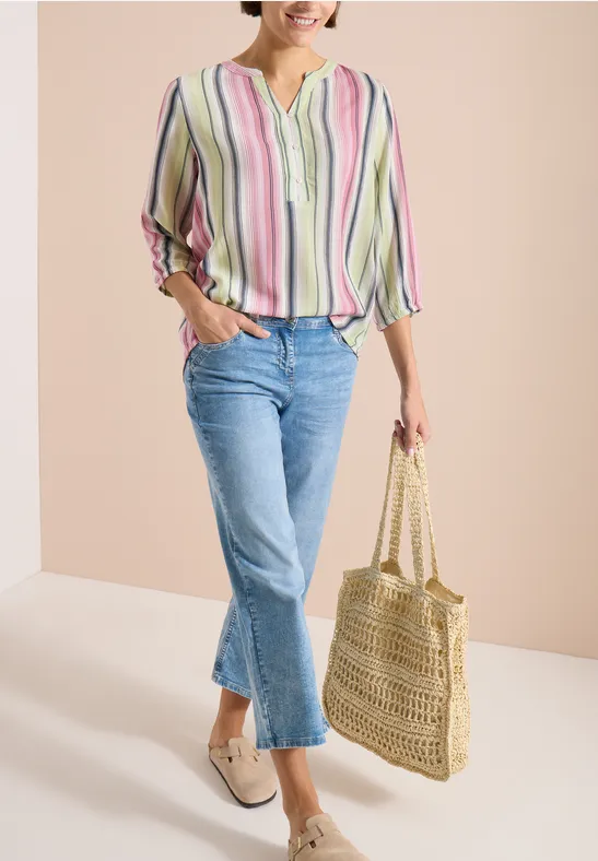 Frau in pastellgestreifter Bluse, blauen Jeans und beigen Slippern hält eine geflochtene Strohtasche.