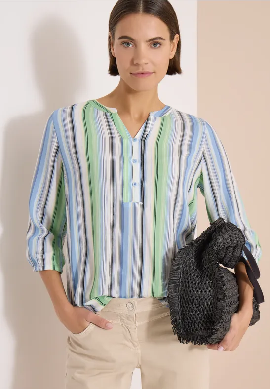 Femme en chemise rayée colorée, pantalon beige et tenant un sac noir tressé.